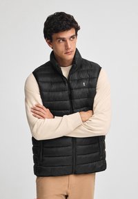 Svart vadderad väst med hög krage, med horisontella quiltade sektioner och en logotypdetalj. Bärs över en ljus beige långärmad skjorta.