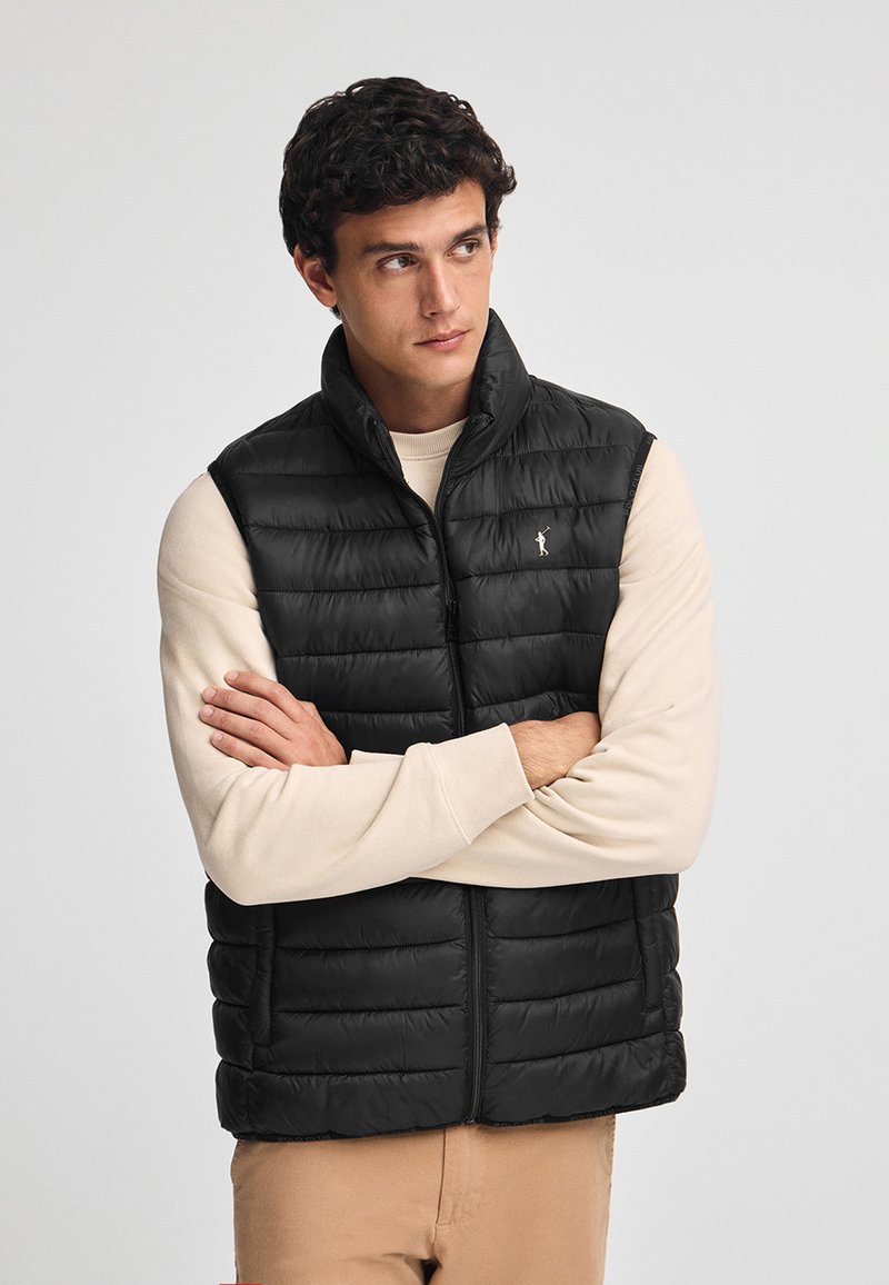 Svart vadderad väst med hög krage, med horisontella quiltade sektioner och en logotypdetalj. Bärs över en ljus beige långärmad skjorta.