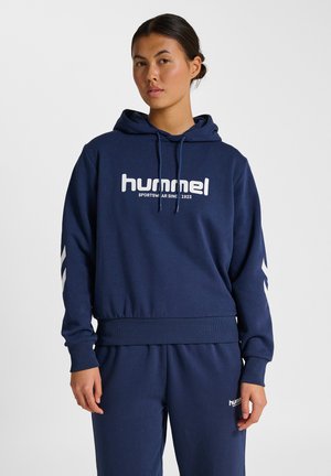 Femme portant un sweat à capuche Hummel bleu marine et un pantalon assorti avec un texte logo blanc, se tenant devant un fond blanc uni.