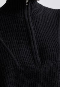 Pull noir en maille côtelée avec un col montant et un détail zippé vertical. La texture est prononcée avec une apparence douce et épaisse.