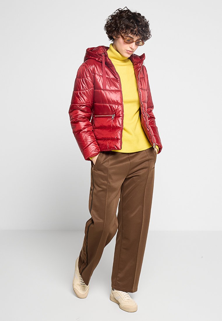 Persoon met krullend haar, die amberkleurige bril draagt, een rode pufferjack, een gele turtleneck, een broek in bruin en beige sneakers, staand voor een witte achtergrond.