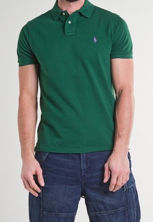 Man die een groen poloshirt met korte mouwen draagt met een klein paars logo op de linkerborst en donkerblauwe cargoshorts.