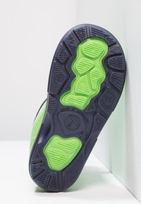 Semelle d'une chaussure avec une base en caoutchouc texturé noir et des accents verts vifs, conçue pour l'adhérence et la durabilité. Fabriquée en Lituanie.