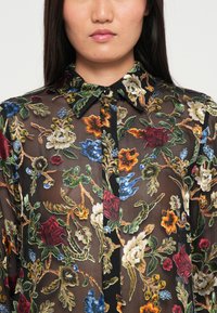 Áttetsző fekete blúz virágos hímzéssel piros, kék, sárga és zöld színekben. Gallér és gombos záródás jellemzi. Texturált anyag élénk mintával.