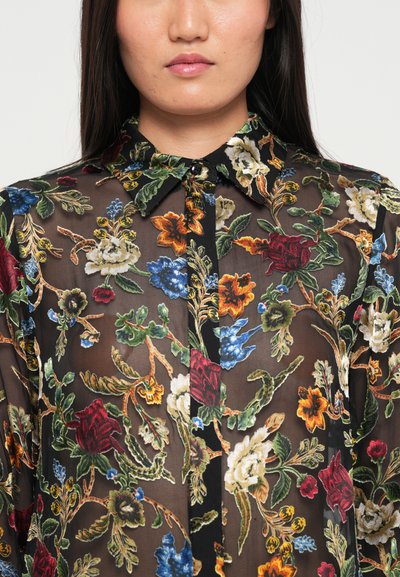 Doorzichtig zwarte blouse met bloemenborduurwerk in rood, blauw, geel en groen. Heeft een kraag en knoopsluiting. Gewaxt stof met een levendig ontwerp.