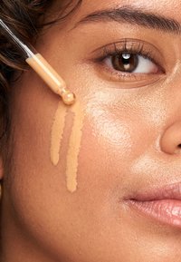 Liquid foundation in a glass dropper is being applied to a cheek. The shade is light beige with a smooth texture against warm, glowing skin.