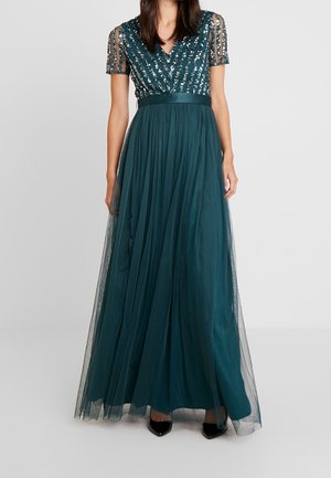 Vrouw draagt een vloerlange donkerblauwe jurk met kralen versierde V-hals en doorschijnende korte mouwen, gecombineerd met zwarte puntige schoenen.