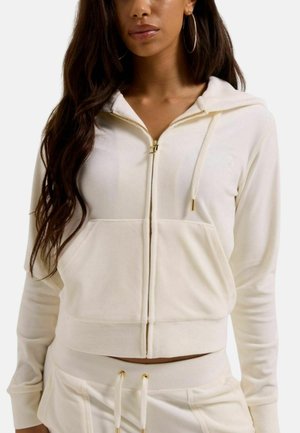 Vrouw draagt een crème kleurige hoodie met gouden rits en bijpassende broek met koordje, staand tegen een effen achtergrond.