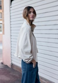 Jeune femme aux cheveux châtain clair portant une veste blanche zippée et un jean bleu, debout de profil avec une main dans la poche près d'un volet fermé.