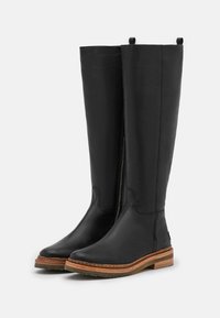 Ein Paar hohe schwarze Lederstiefel mit braun genähten Sohlen und seitlichen Reißverschlüssen, die auf einem weißen Hintergrund aufrecht stehen.