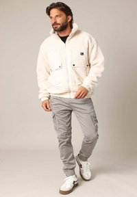 Veste en polaire blanche avec deux poches frontales, pantalon cargo gris et baskets blanches avec des accents noirs. Le modèle se tient sur un fond neutre.