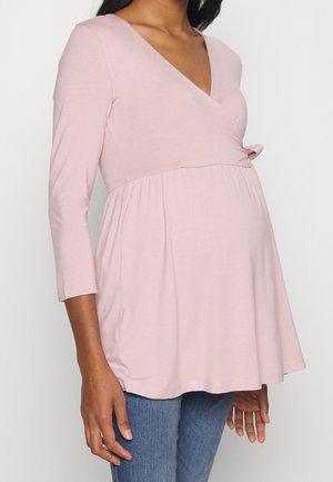 Femme enceinte portant un haut de maternité rose à manches longues avec col en V et un jean bleu, debout devant un fond uni.