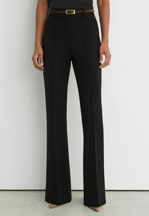 Vrouw draagt zwarte, op maat gemaakte broek met hoge taille en plooien aan de voorkant, bruine riem en beige puntige schoenen, staand tegen een witte muur.
