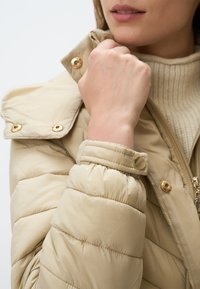 Beige pufferjack met een hoge kraag, gouden hardware-accenten en gestikte secties. De mouw is gathered bij de manchetten.