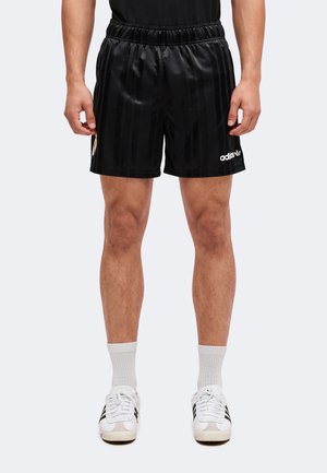 Uomo con pantaloncini sportivi neri Adidas, calzini bianchi e scarpe da ginnastica bianche, in piedi con le mani rilassate lungo i fianchi.