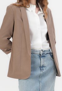 Mujer vestida con una camisa blanca de botones, un blazer beige y una falda de mezclilla azul clara, con las manos en los bolsillos del blazer, contra un fondo blanco.