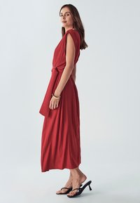 Červené šaty po kolena se zavázaným pasem, krátkými rukávy a měkkou, texturovanou látkou. Nosené s černými sandály na podpatku.