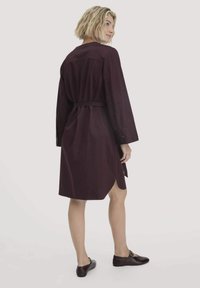 Frau mit kurzen blonden Haaren, die ein dunkelviolettes, tailliertes Kleid und dunkle Flache trägt, steht und schaut über ihre Schulter vor einem schlichten Hintergrund.