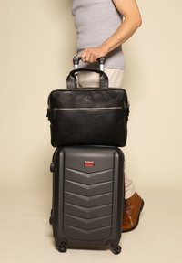 Valigetta nera in pelle con una tasca con zip sopra una valigia rigida grigia, caratterizzata da un motivo a zigzag e ruote girevoli. Scarpe marroni visibili.