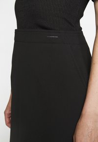 Schwarzer taillierter Rock mit glatter Textur, Slim Fit und dezenten Seitentaschen. Enthält ein kleines Markenakzent an der Taille.