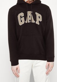 Felpa nera con tasca a marsupio, caratterizzata da un grande logo "GAP" color beige. Presenta lacci e polsini a coste sulle maniche.
