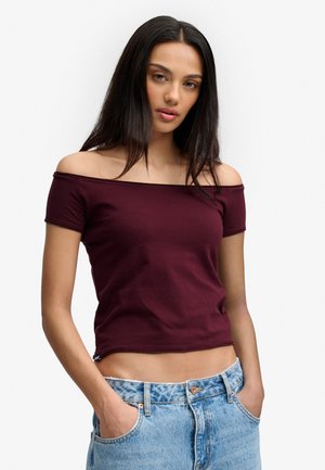 Jonge vrouw die een bordeauxrode off-shoulder top en lichtblauwe spijkerbroek draagt, met handen in de zakken, staand tegen een effen witte achtergrond.