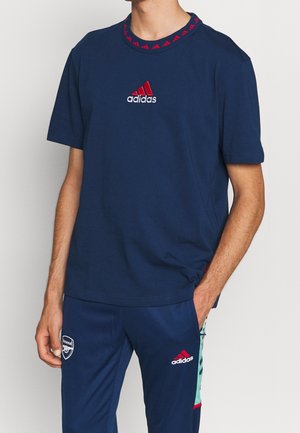 Homme portant un t-shirt Adidas bleu marine et un pantalon de survêtement assorti avec le logo du club de football Arsenal, debout avec une main dans la poche.