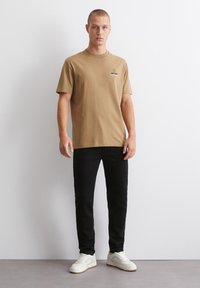 Brun bomull t-skjorte med korte ermer og rund hals, kombinert med sorte jeans og hvite joggesko, stående mot en ensfarget bakgrunn.
