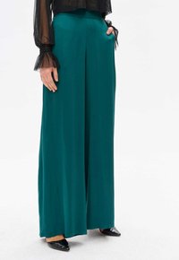Turkusowe spodnie o szerokich nogawkach, wykonane z gładkiego materiału, z wysokim stanem i bocznymi kieszeniami, połączone z czarnymi butami na szpilkach.