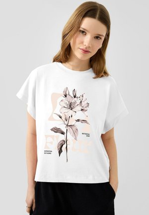 Junge Frau, die ein weißes T-Shirt mit einer großen detaillierten Blumenillustration und Text "Exposition de Fleurs" und "Botanical Soir." trägt