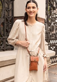 Smeđa kožna torba preko tijela s preklopom i kopčom, nosi se uz svijetlosmeđu haljinu na dugme s remenom i napuhanim rukavima.