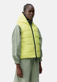 Gul puffer vest med hette, med en frontglidelås, kombinert med en lys grønn, løs passende antrekk og elastiske mansjetter.