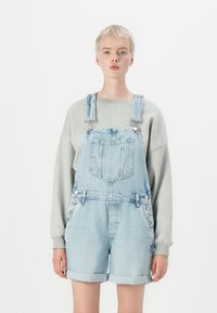 Salopette in denim azzurro chiaro con bretelle regolabili, tasca frontale e orlo arrotolato. Abbinata a una felpa grigia a maniche lunghe.
