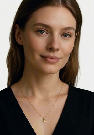 Junge Frau mit heller Haut und glattem braunem Haar trägt ein schwarzes Oberteil und eine goldene Halskette mit einem "E"-Anhänger, neutraler Hintergrund.