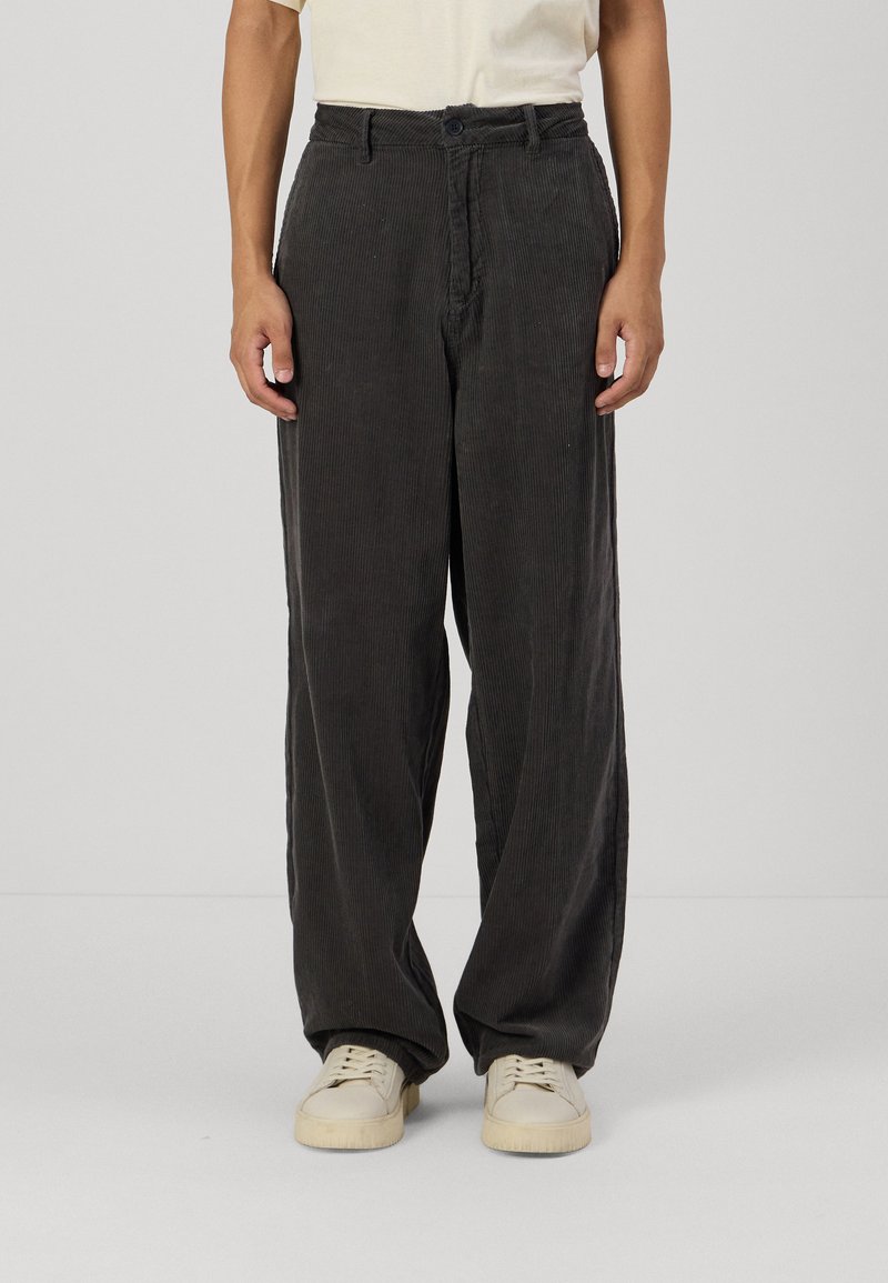 Pantalones de pana en gris oscuro con un diseño de pierna ancha, con cierre de botón y tela texturizada, combinados con zapatillas de deporte en blanco roto.