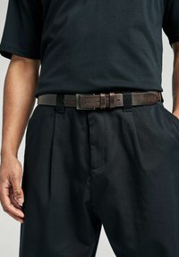 Ceinture en cuir marron avec une surface texturée, dotée d'une boucle en métal argenté et de plusieurs trous d'ajustement, portée sur un pantalon noir.