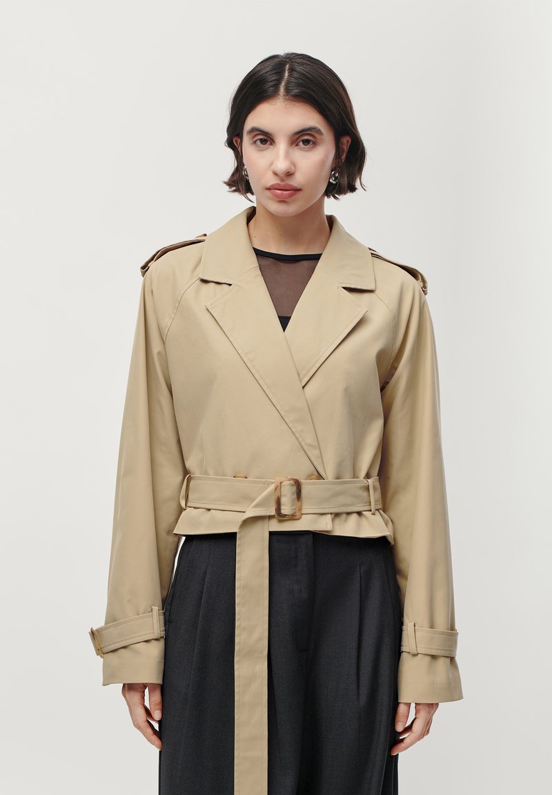 Beige trenchcoat met een wikkelontwerp, brede revers, een ceintuur in de taille en lange mouwen. Bevat schouderepauletten en een gladde textuur.