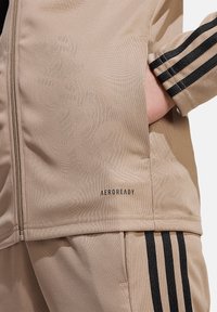 Beige Sportjacke mit Reißverschluss, strukturiertem Stoff und schwarzen Streifen an den Ärmeln. "AEROREADY"-Logo nahe dem Saum.