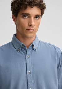Lichtblauw overhemd met knopen van een gestructureerde stof, voorzien van een kraag, een placket met twee knopen en een subtiel geribbeld patroon.