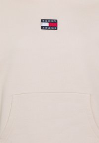 Krémová mikina s přední kapsou a černou, červenou a bílou vyšívanou nášivkou s nápisem "TOMMY JEANS."
