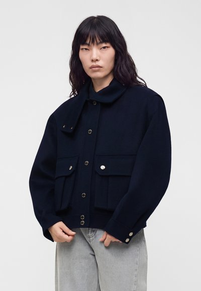 Personne aux longs cheveux foncés portant une veste boutonnée bleu marine foncé avec de grandes poches et un pantalon gris clair sur un fond uni.