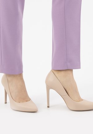 Tacchi a stiletto beige con una texture liscia e punta affusolata, abbinati a pantaloni su misura di un leggero colore viola, che mostrano un look contemporaneo.