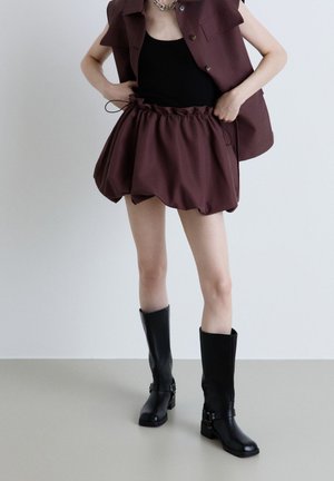 Persona che sistema una gonna marrone arricciata abbinata a un gilet coordinato, top nero e stivali neri al ginocchio su sfondo semplice.