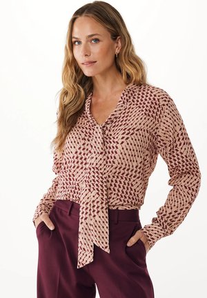 Blusa a maniche lunghe di un rosa chiaro con un motivo geometrico borgogna. Presenta un collo annodato e un tessuto morbido e testurizzato. Abbinata a pantaloni di un marrone scuro.