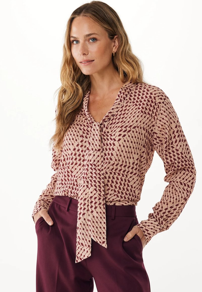 Blusa a maniche lunghe di un rosa chiaro con un motivo geometrico borgogna. Presenta un collo annodato e un tessuto morbido e testurizzato. Abbinata a pantaloni di un marrone scuro.