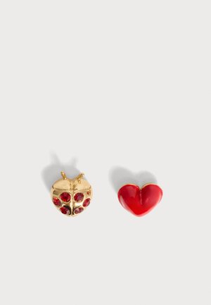Clou d'oreille coccinelle en or avec des pierres rouges et un clou en forme de cœur rouge brillant sur un fond clair uni.