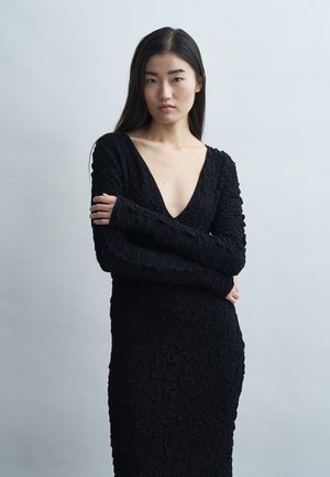 Frau mit langen dunklen Haaren, die ein strukturiertes schwarzes Kleid mit tiefem V-Ausschnitt trägt, die Arme verschränkt, und vor einem schlichten hellgrauen Hintergrund steht.