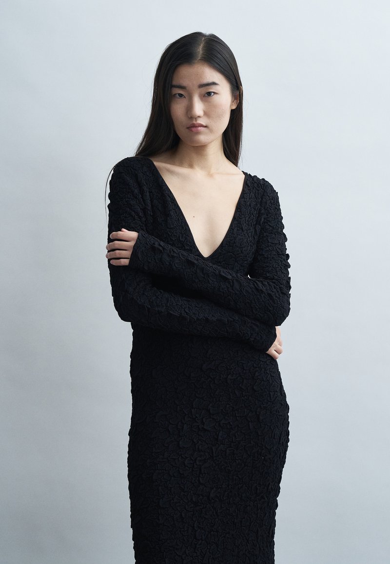 Frau mit langen dunklen Haaren, die ein strukturiertes schwarzes Kleid mit tiefem V-Ausschnitt trägt, die Arme verschränkt, und vor einem schlichten hellgrauen Hintergrund steht.