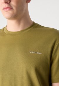 Tricou din bumbac verde măsliniu cu guler rotund și un logo brodat alb "Calvin Klein" pe piept. Textură netedă, mâneci scurte.