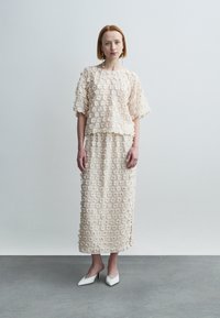 Ensemble deux pièces beige clair comprenant un haut à manches courtes et une longue jupe, ornés d'appliqués floraux, accompagné de talons blancs.